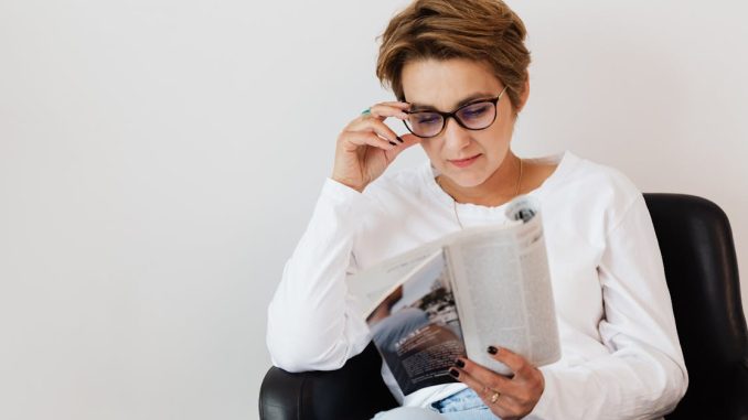 positive-adult-short-haired-female-in-casual-wear-and-eyeglasses-reading-interesting-magazine-while